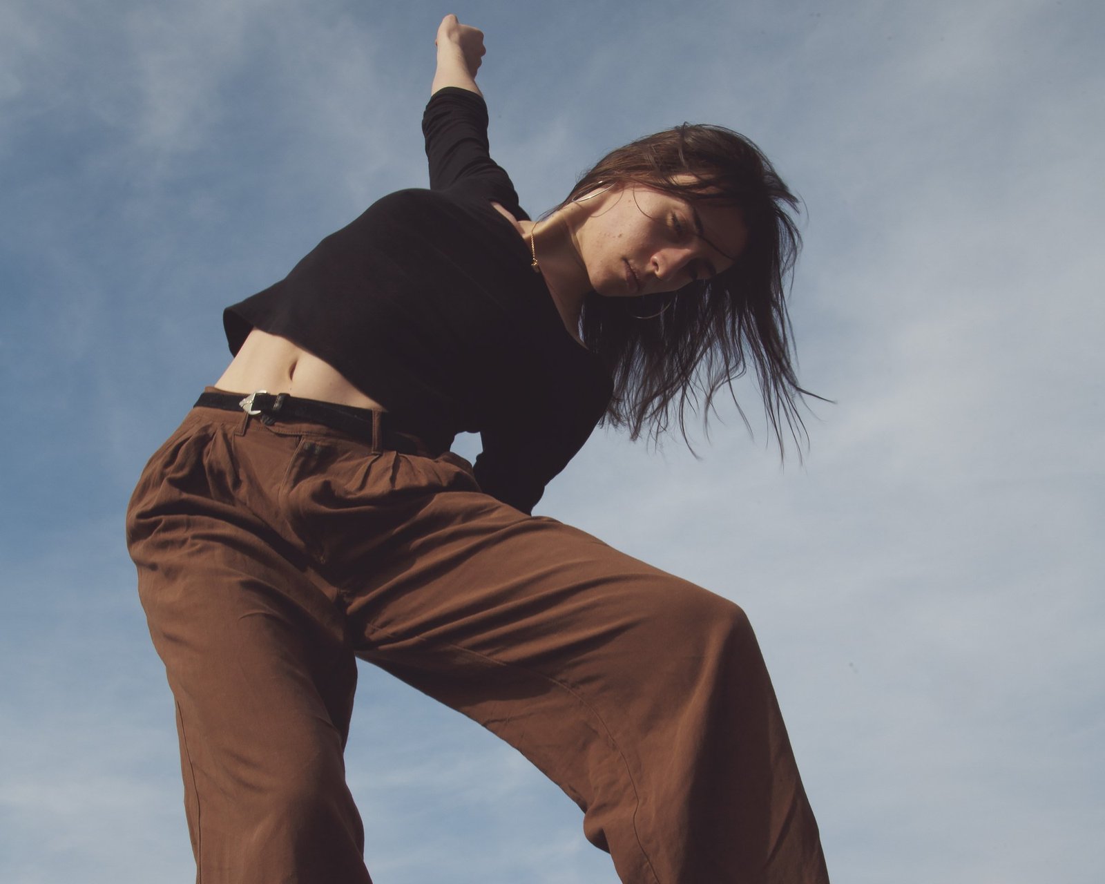 A dancer in brown trousers and a black crop top leaning back, arm raised against cloud.
