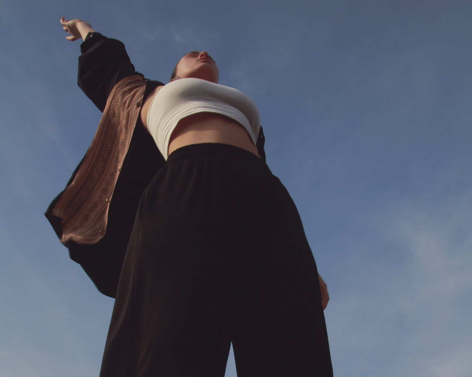 A dancer in a white crop top and dark jacket, chin lifted, one arm extended into the sky.