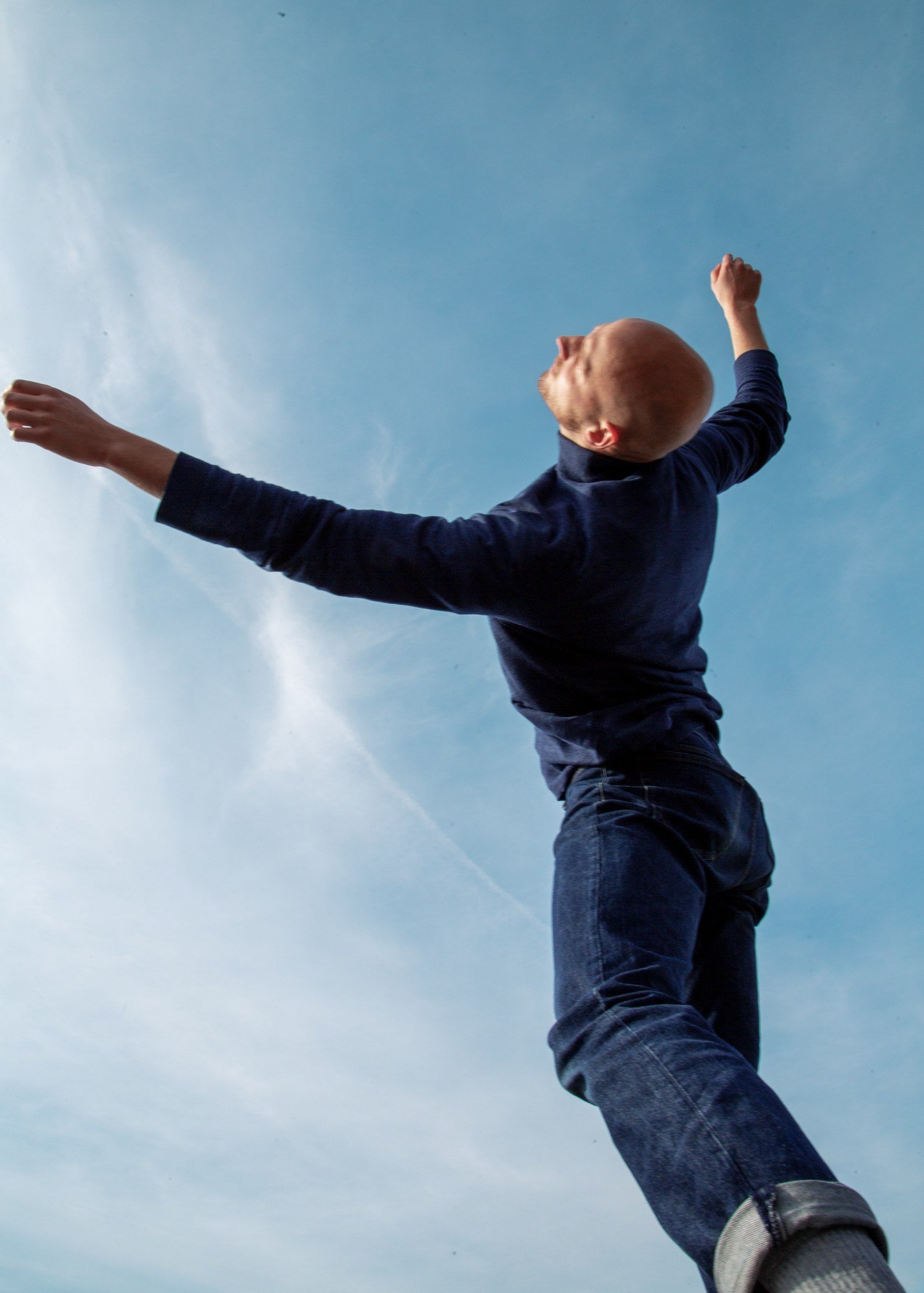 A figure in navy with both arms flung wide, head thrown back against pale sky.