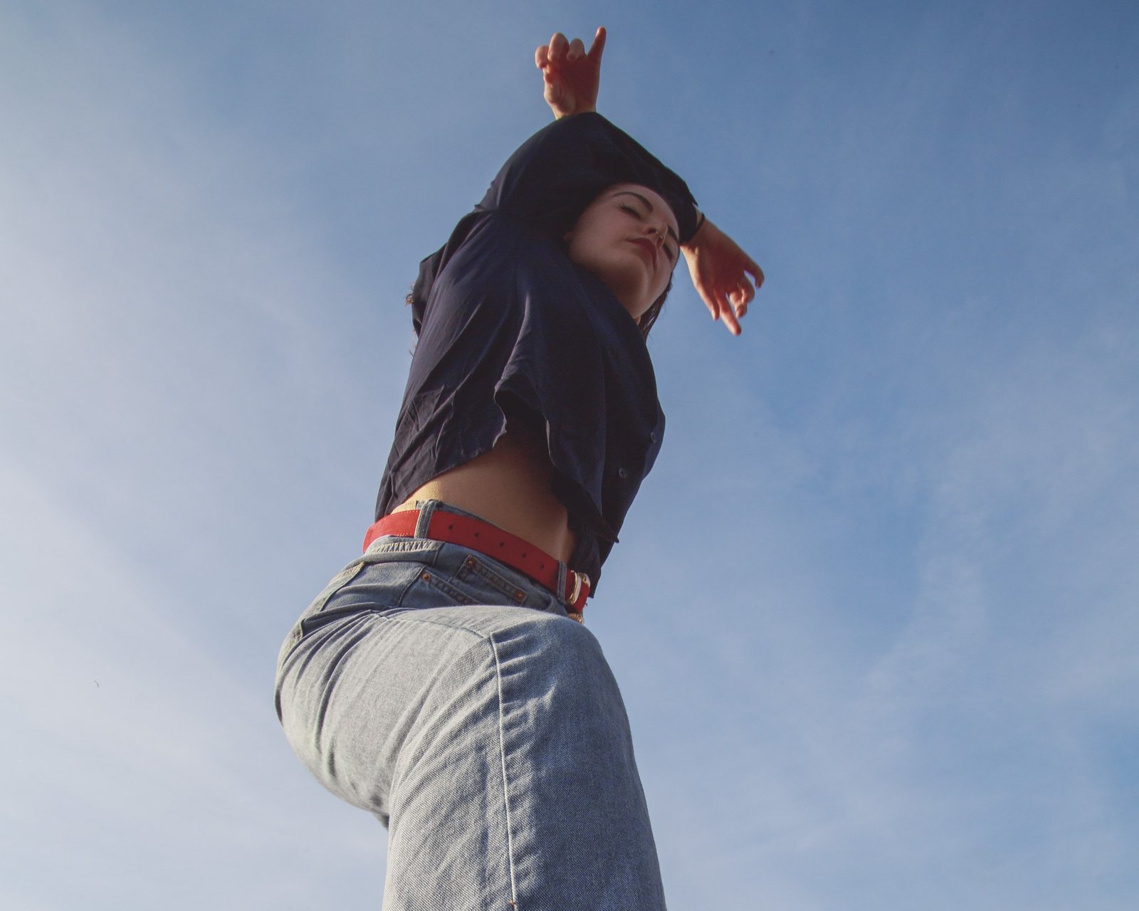 A dancer in light denim and a navy shirt, both arms crossed above the head, eyes closed.