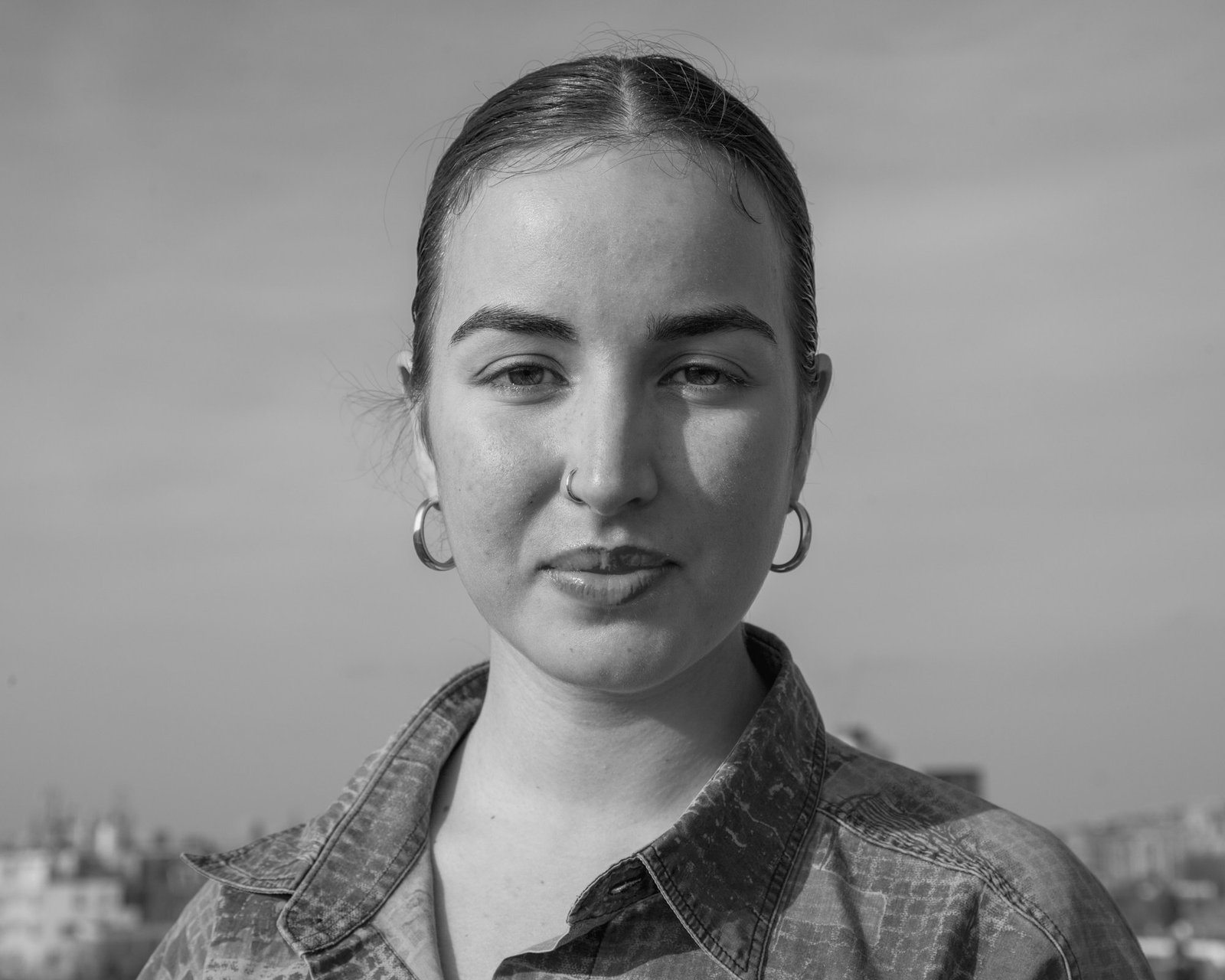 A woman with slicked-back hair and hoop earrings in a checked shirt, city roofs behind her.