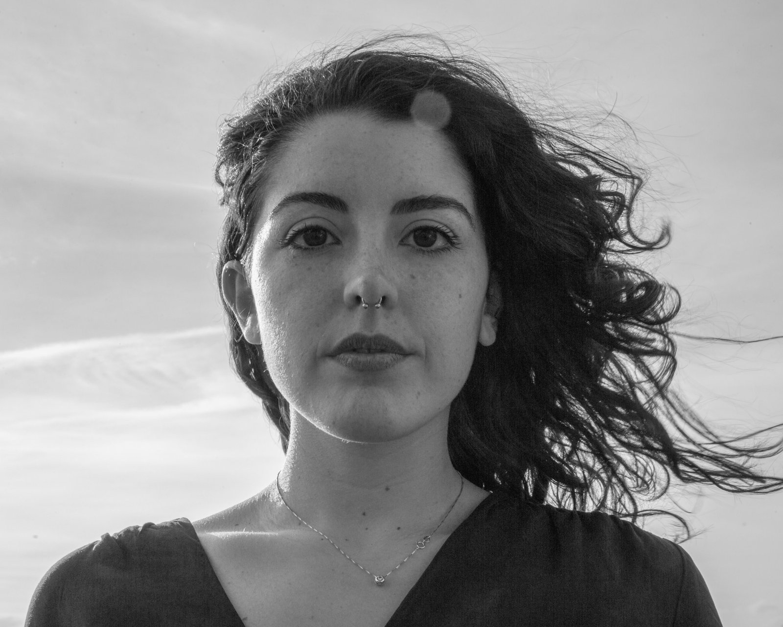 A woman with dark hair blown loose across a bright sky, looking straight into the lens.