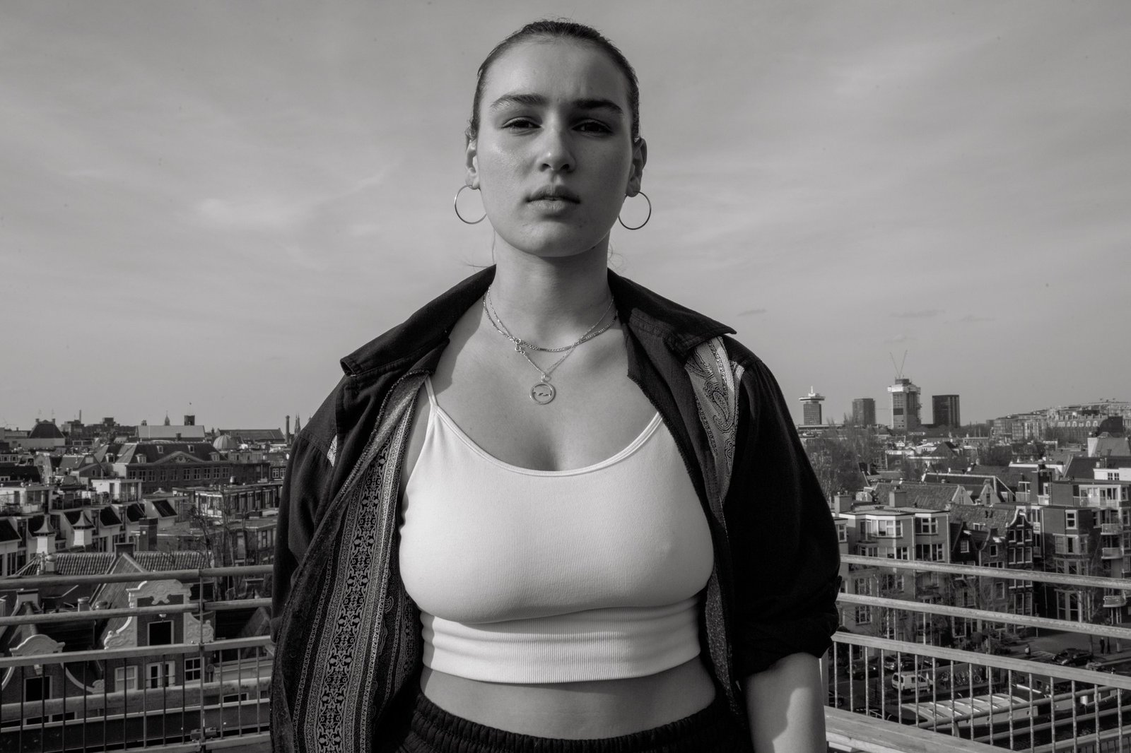 A woman in a white top and open leather jacket on a rooftop, Amsterdam spread out behind.