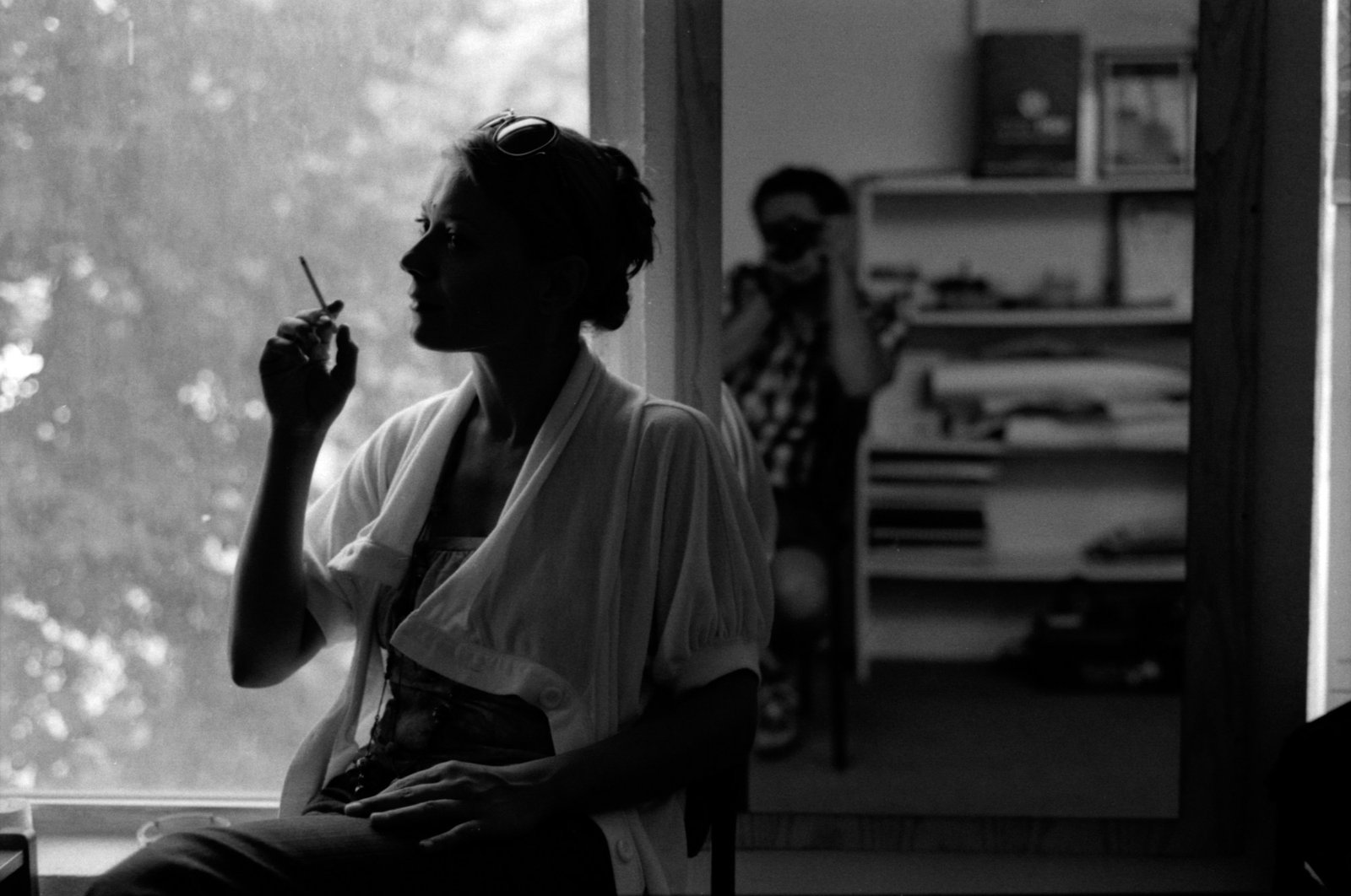 A silhouetted figure smoking at a window beside a second frame of the room behind.