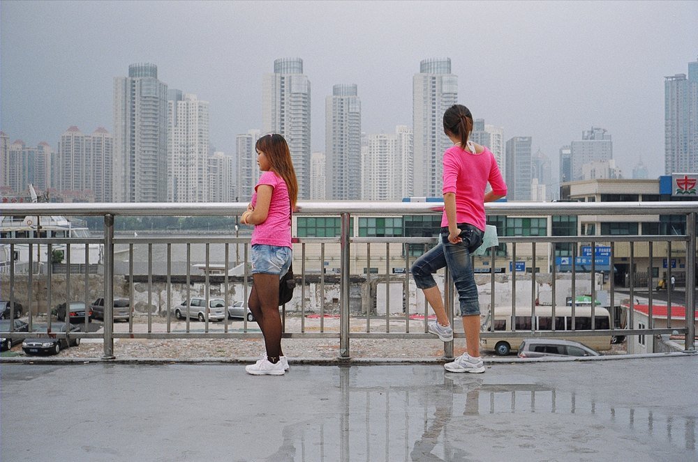 Two young women in pink at the railing of a raised walkway, a hazy skyline of high-rise towers behind them.