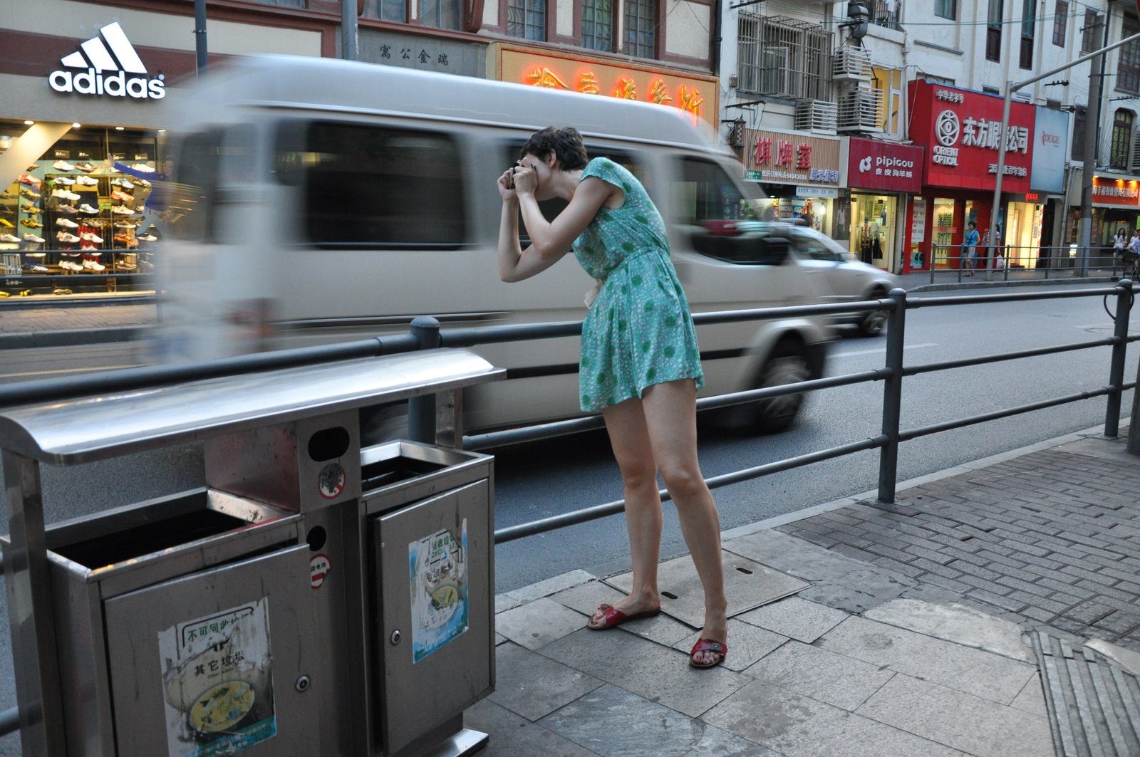 A woman bent over her camera on a busy pavement as a white van streaks past behind her.