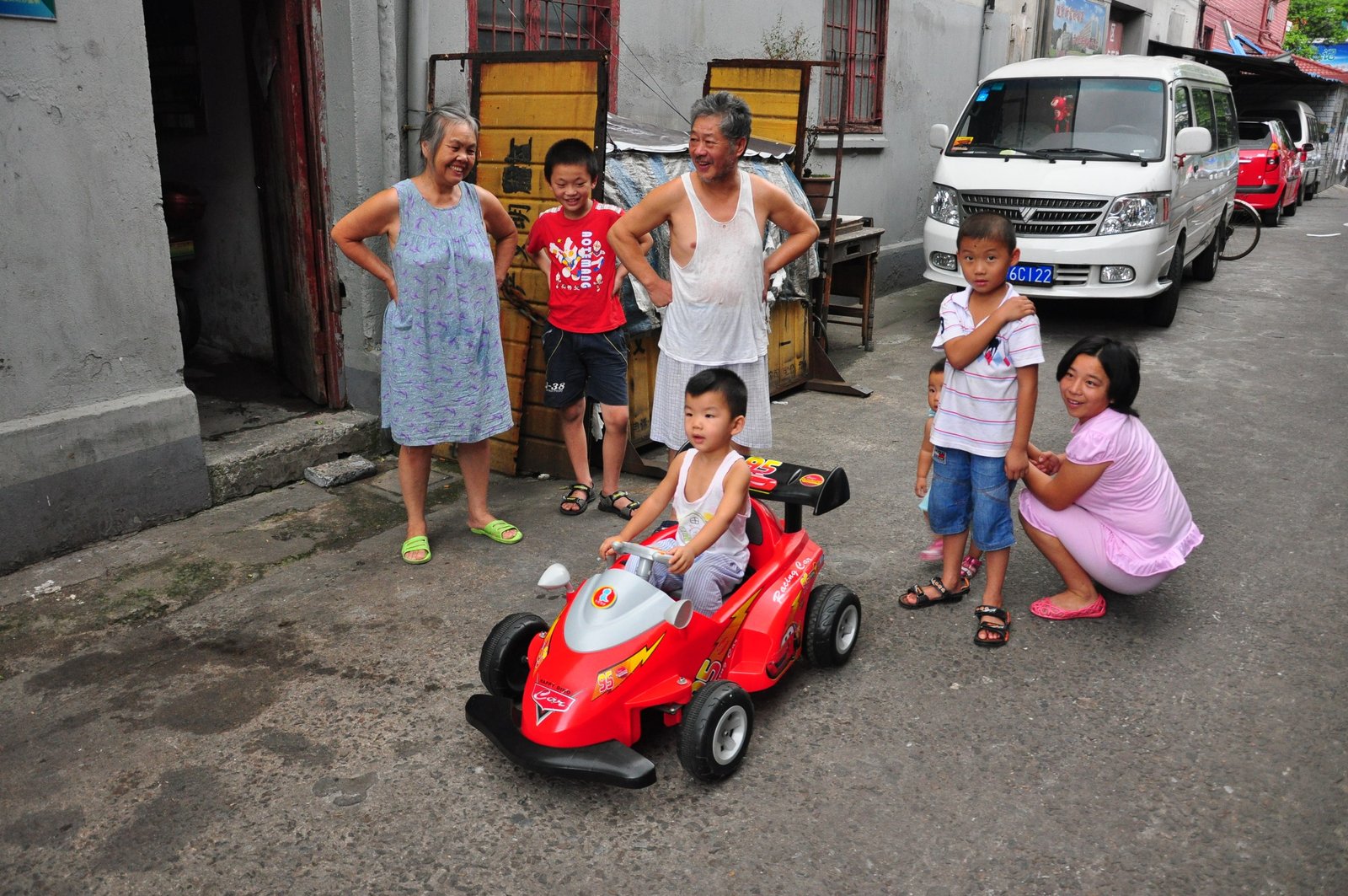 A family gathered in an alley around a small child on a red toy car, others watching from the kerb.