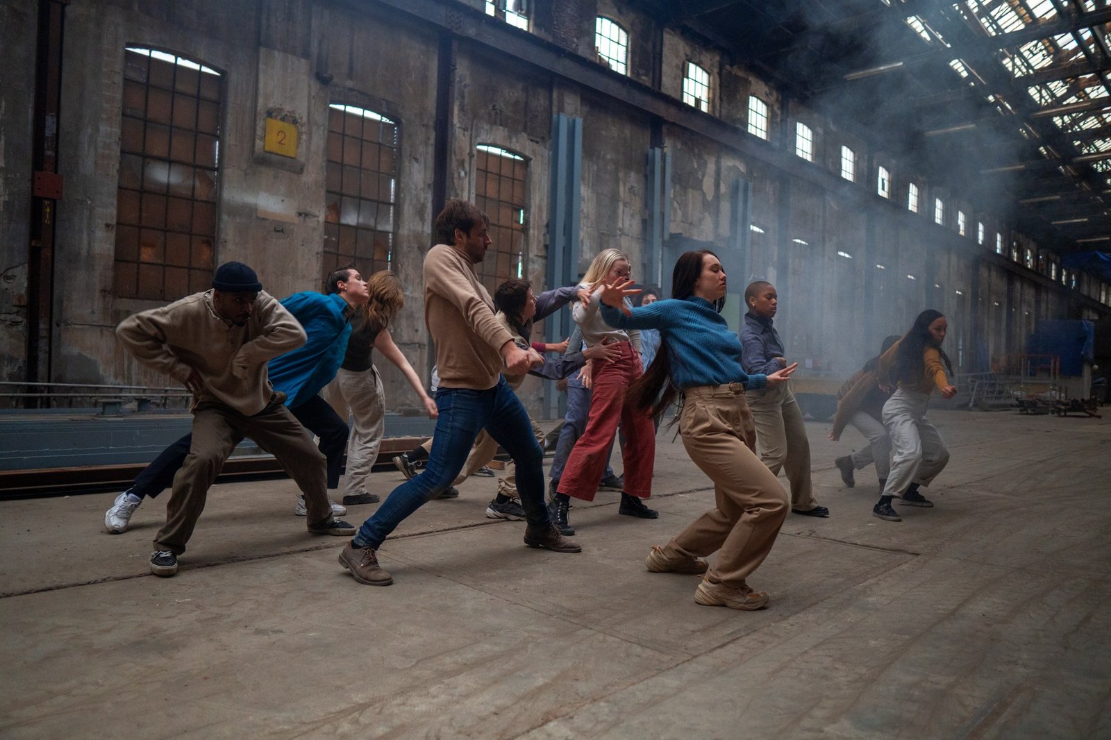 A line of dancers braced mid-phrase down the length of a long factory hall, tall windows either side.