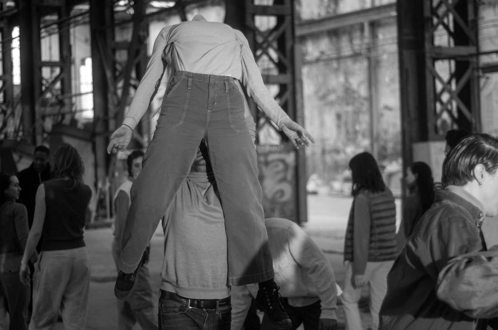 A figure caught mid-jump, head lost inside a pulled-up shirt, onlookers along an industrial wall.