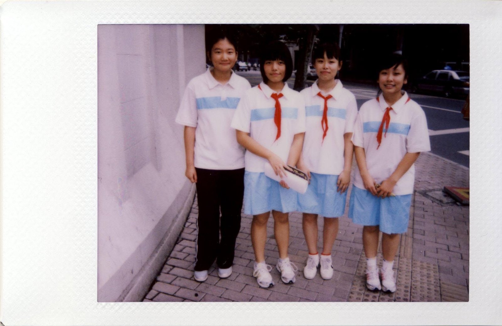 Four schoolgirls in white and pale blue uniforms with red neckerchiefs on a pavement.