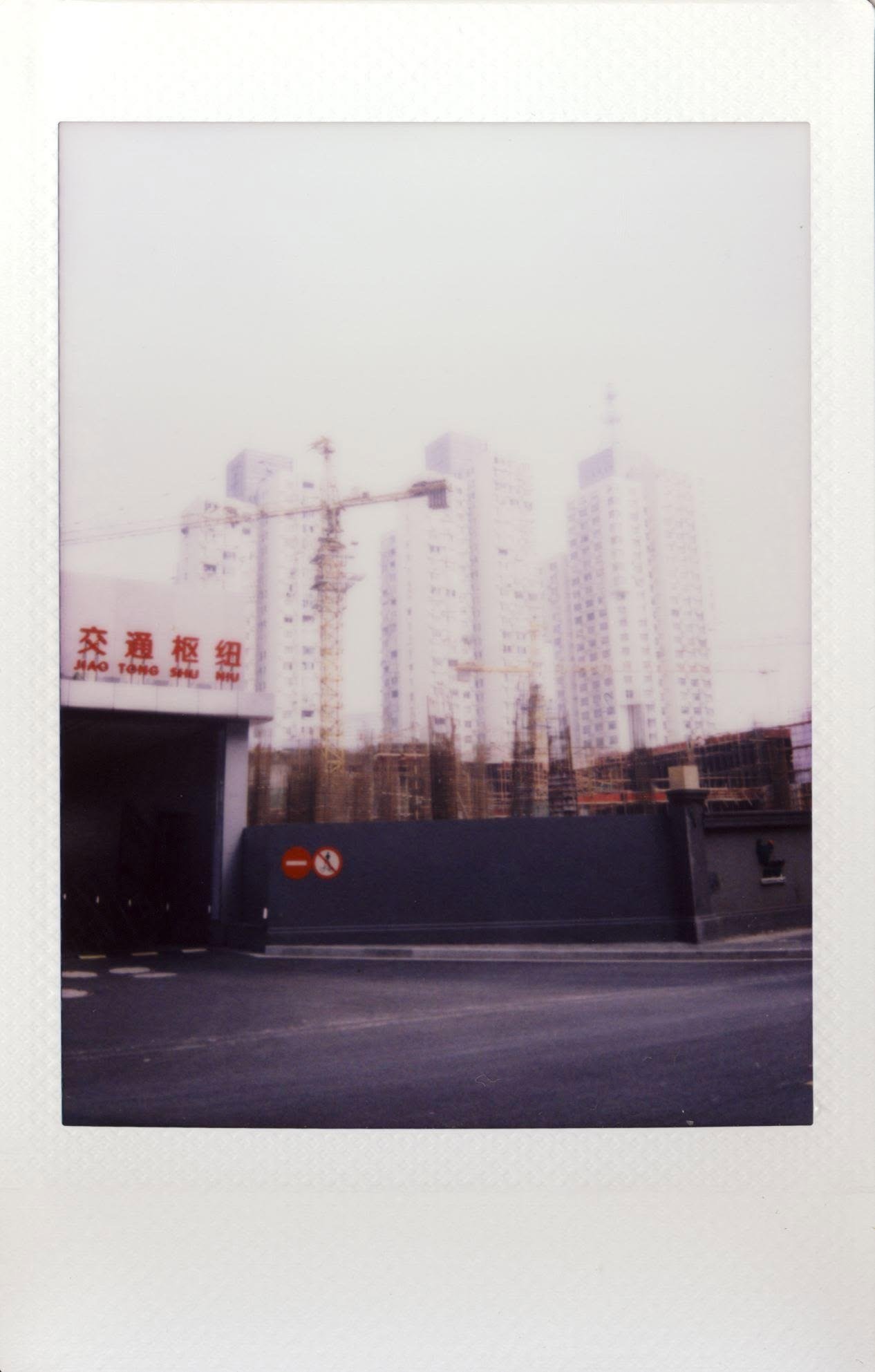 Apartment towers under construction behind a blue hoarding, cranes above, a red transport-hub sign.