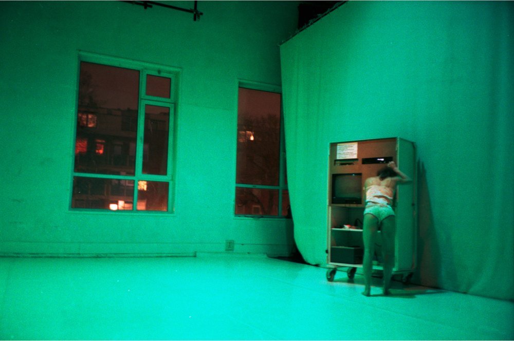 A room flooded with green light, a green cyclorama curtain, a small figure crouched by a cabinet.