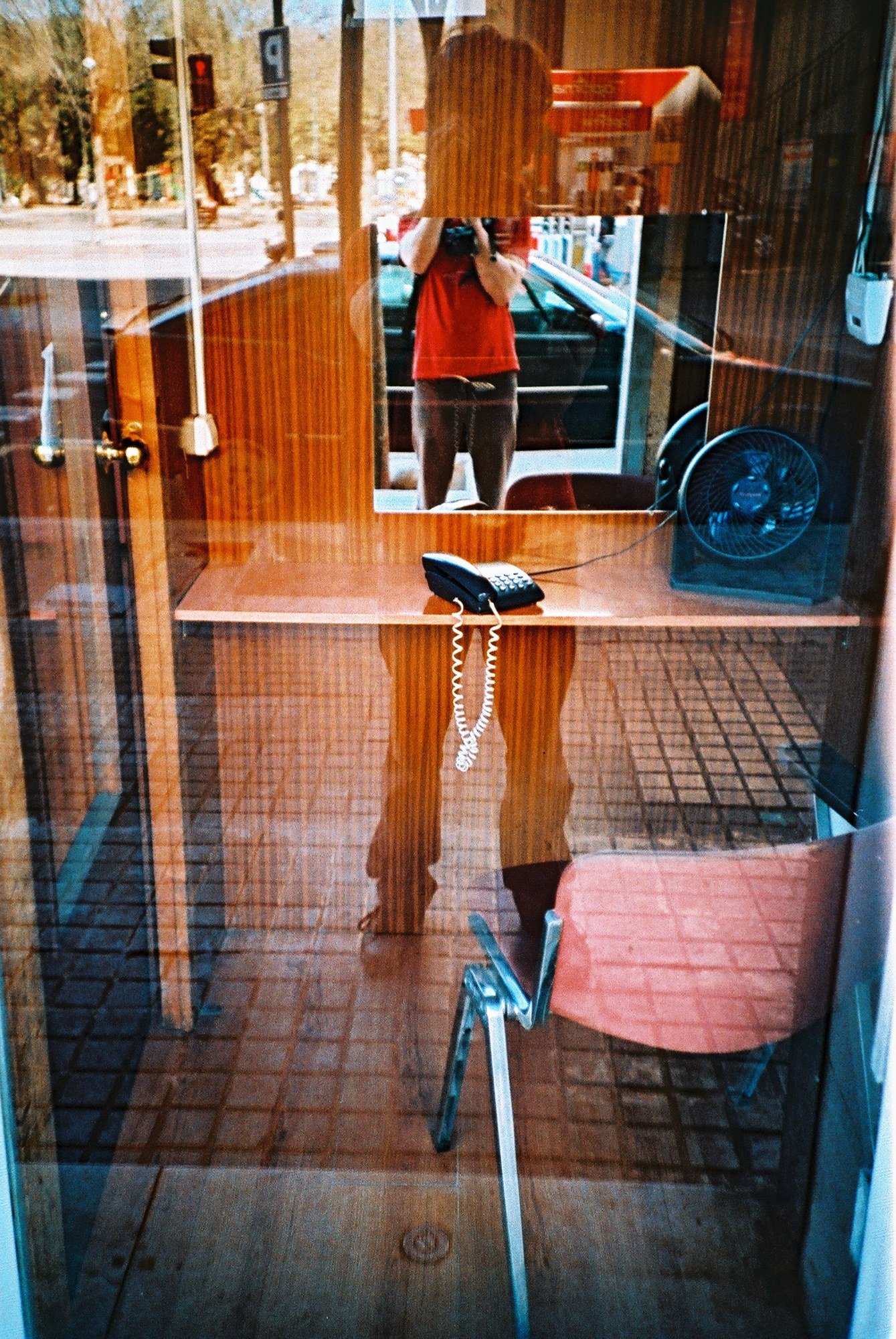 An empty reception booth seen through glass — wooden desk, corded telephone, salmon-pink chair — with the photographer reflected in the mirror above it, camera raised.