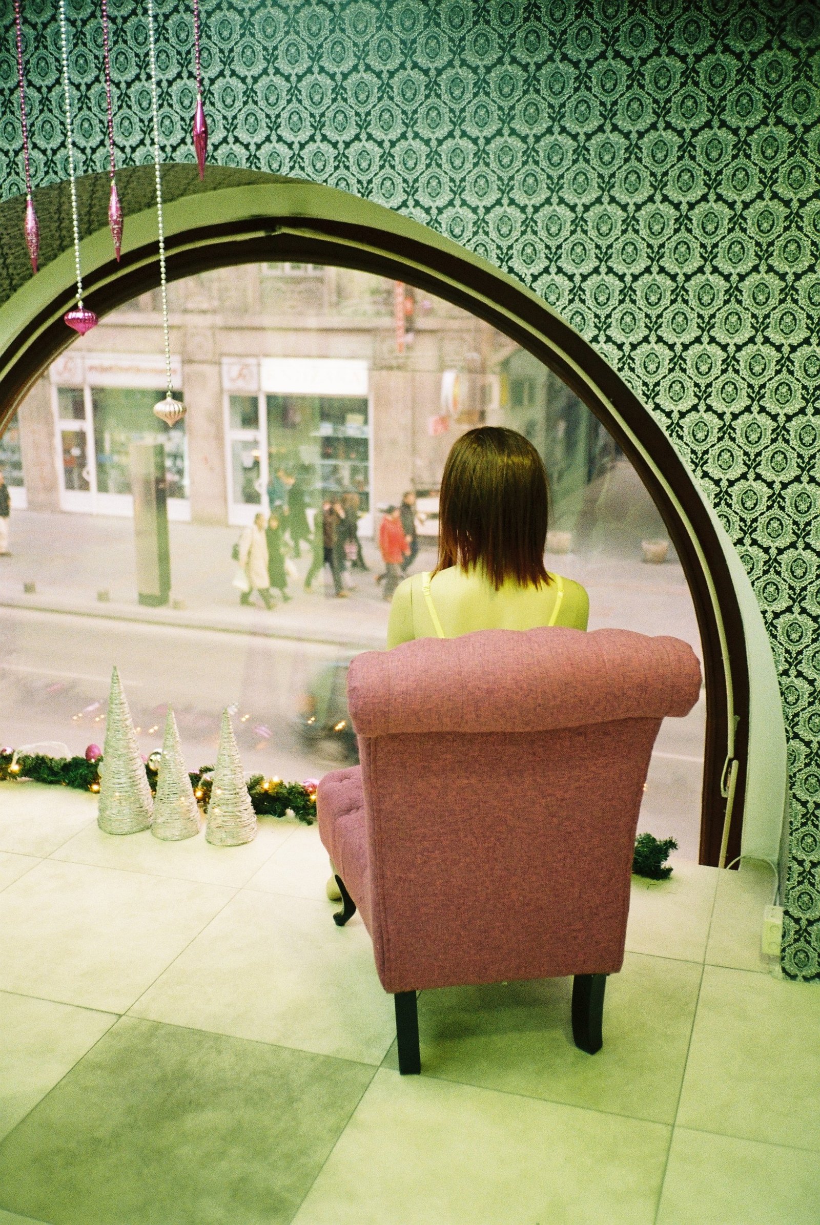 A room papered in green damask, a round mirror above a pink armchair, a woman seated with her back to the camera and the street reflected behind her.