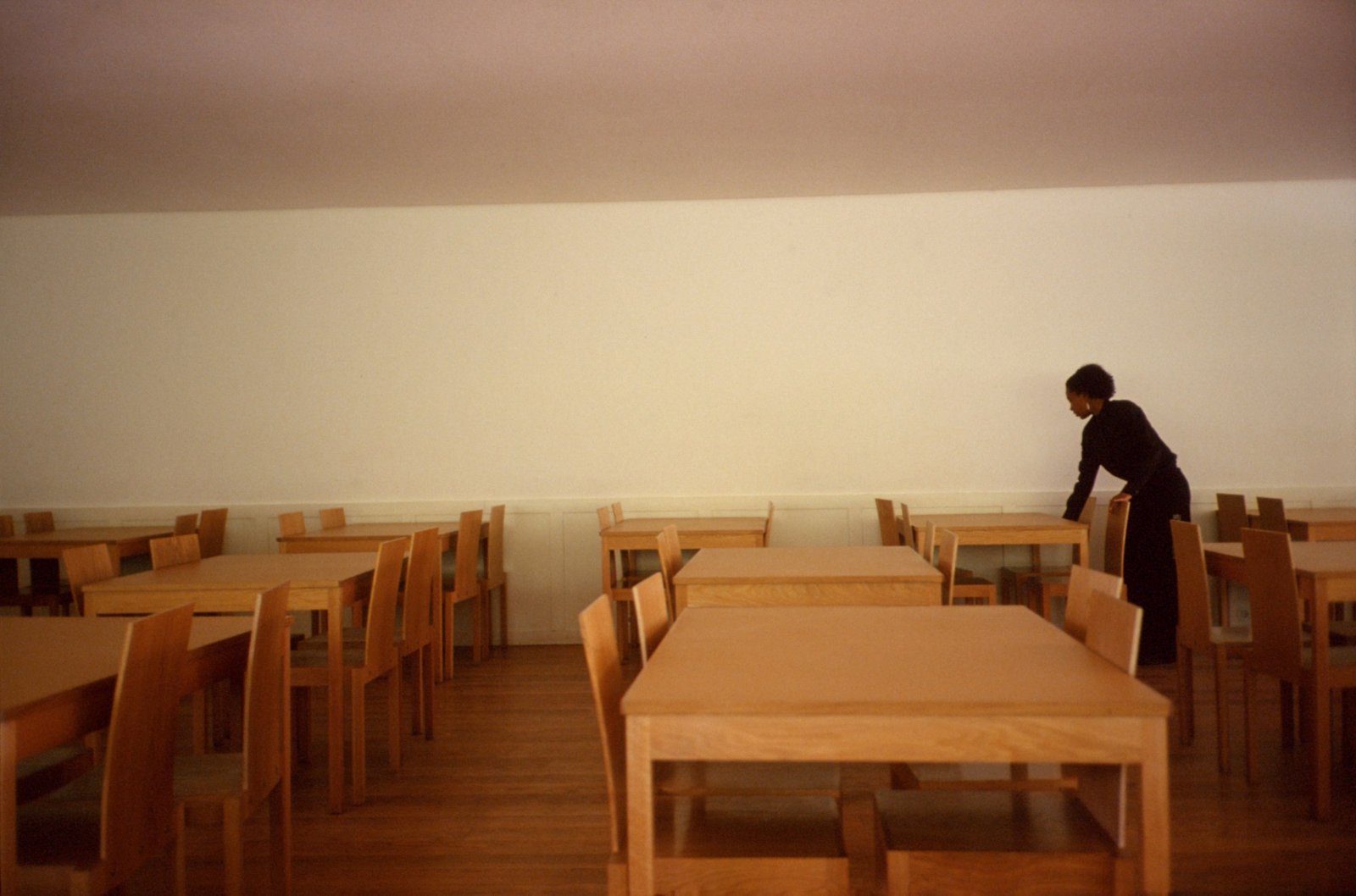 An empty canteen in warm afternoon light, long wooden tables, one figure at the far wall.
