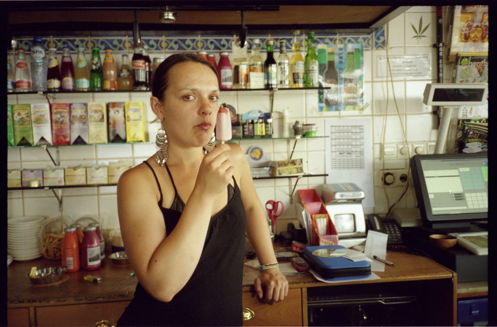 A woman behind a bar counter surrounded by bottles and shelves, looking at the camera.